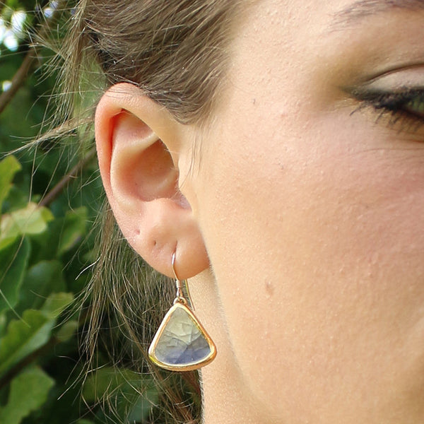 Close-up of a person wearing a gold earring with a blue gemstone against a blurred green background